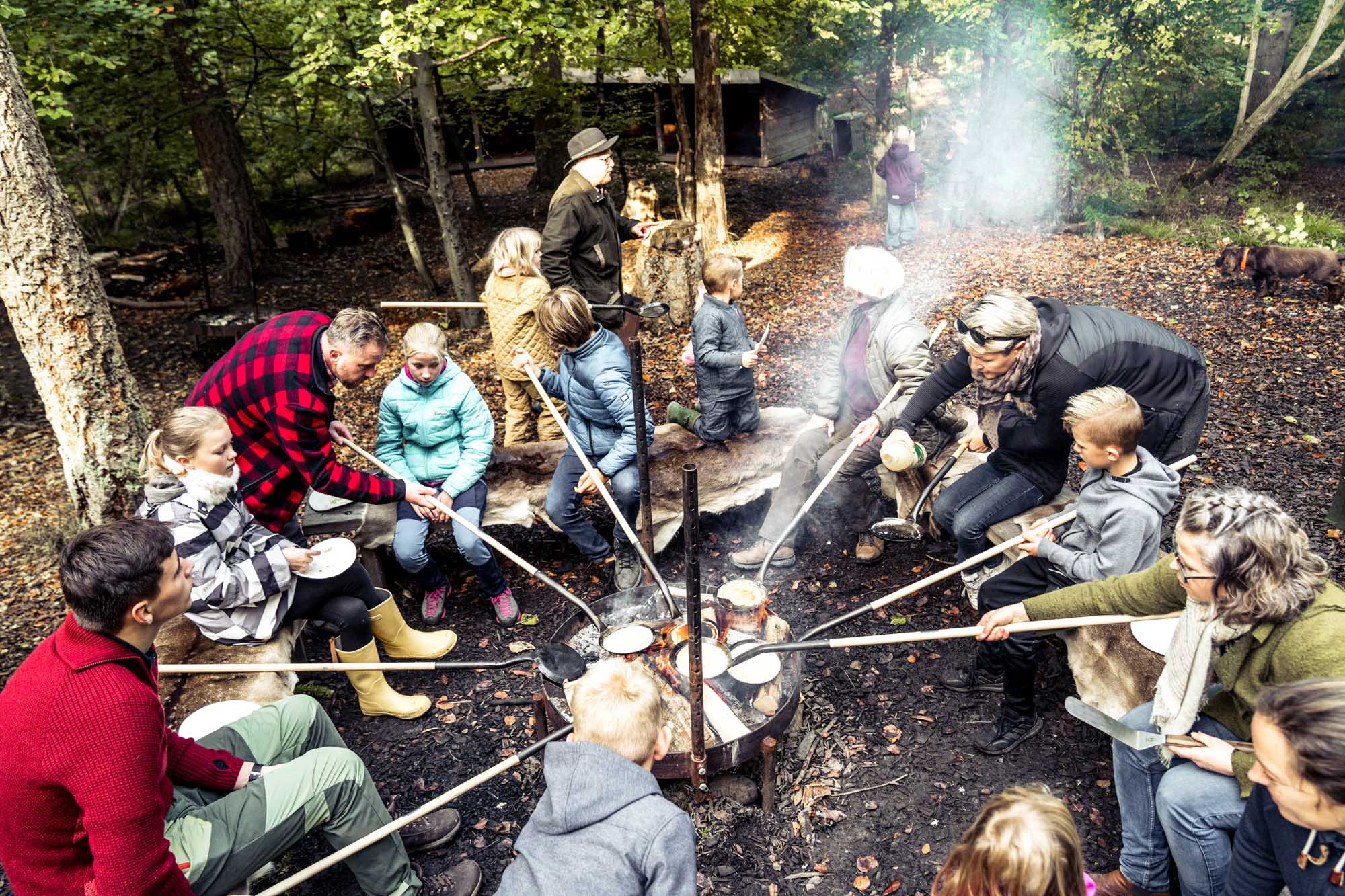 En gruppe børn og vokse sidder om et bål på en shelterplads og laver pandekager 